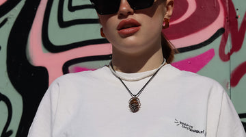 Person looking down at the camera with sunglasses and a white t-shirt on. She is standing in front of a graffiti'd background.